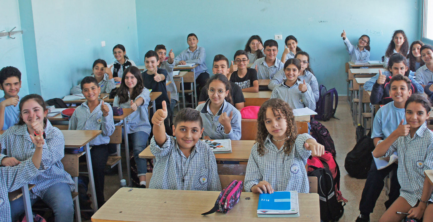 Schulkinder aus dem Libanon sitzen in hellblauer Schuluniform in einem Klassenzimmer an Schultischen. Sie lachen in die Kamera, einige von ihnen strecken ihren Daumen nach oben.