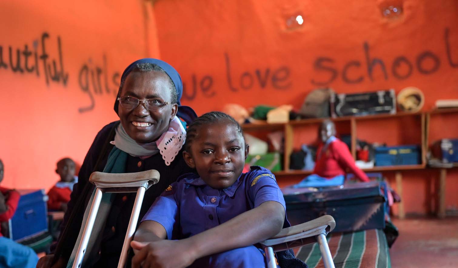 In einem Klassenzimmer mit rot-orangenen Wänden ist im Vordergrund eine Ordensschwester aus Kenia zu sehen, die neben einem Mädchen mit Behinderung sitzt. Vor der Ordensschwester lehnt die Krücke des Mädchens.