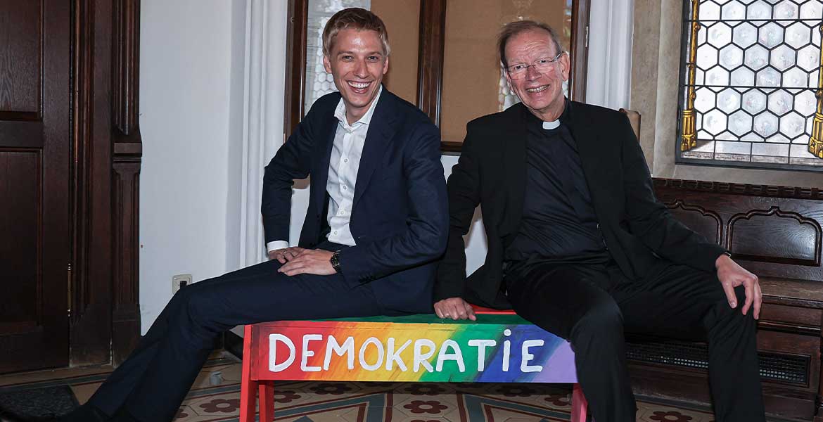 Münchens Oberbürgermeister Dominik Krause (links) sitzt mit missio-Präsident Huber auf einer Bank mit der Aufschrift "Demokratie".".