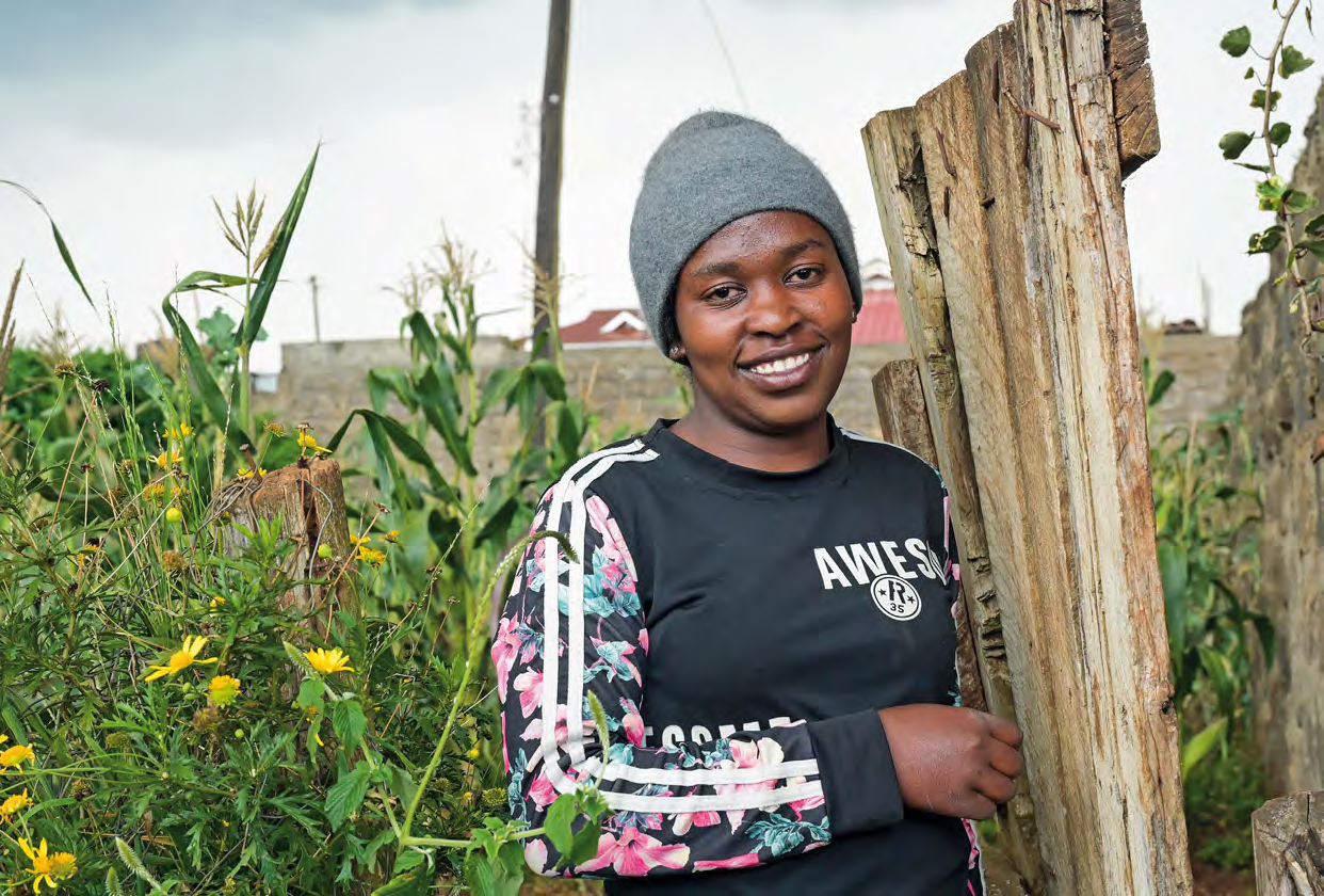 Eine junge Frau mit grauer Mütze steht in ihrem Garten an einen improvisierten Zaun gelehnt und lächelt.