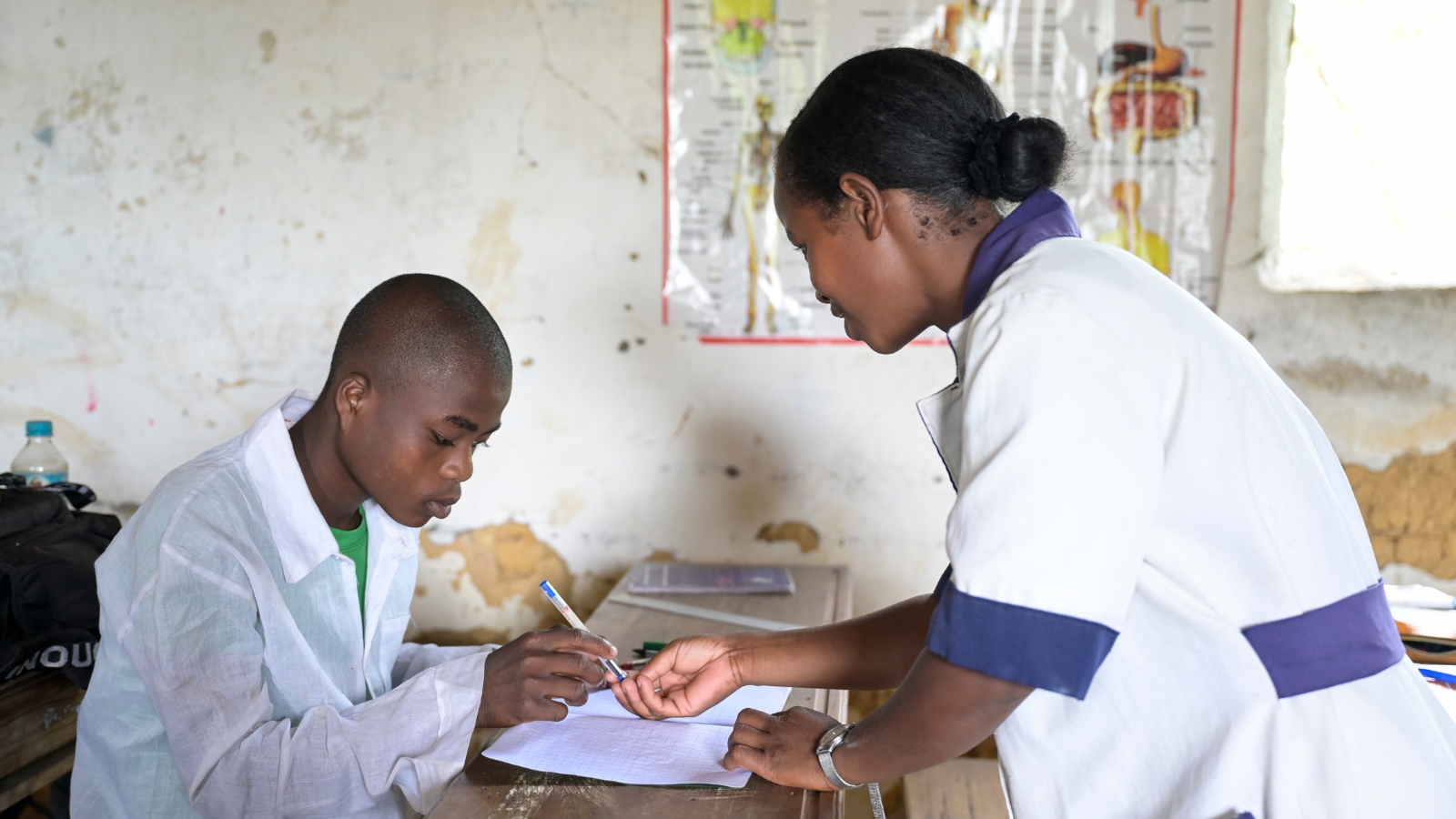 Ein jugendlicher Schüler in weißem Hemd beugt sich über ein Schulheft. Die Lehrerin wendet sich ihm zu, um ihm etwas zu zeigen.