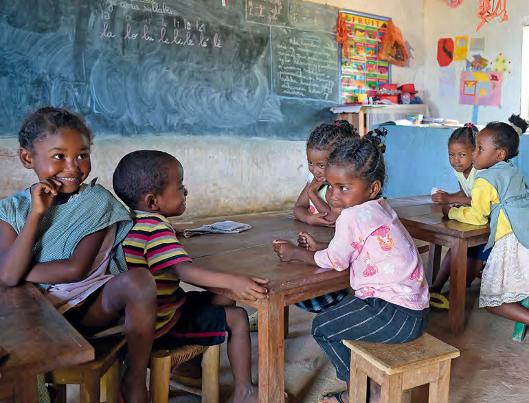 Einfacher Klassenraum mit gestampftem Lehmboden. Grundschulkinder sitzen an kleinen Tischen vor der Tafel.