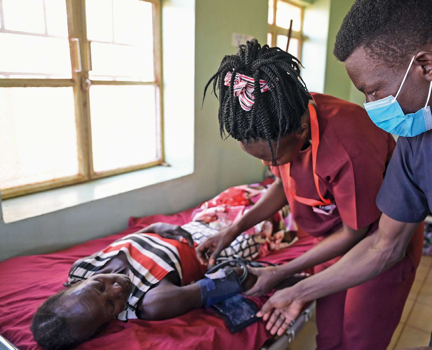 Edia Atanasio steht am Bett einer Patientin und legt ihr das Blutdruckmessegrät an. Ein Arzt oder Krankenpfleger steht neben ihr und gibt ihr Tipps.