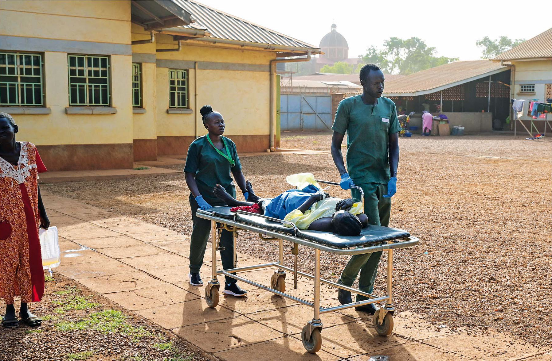 Eine Patientin wird vom Krankenhauspersonal auf einer Liege über den Hof des Klinikgeländes transportiert.