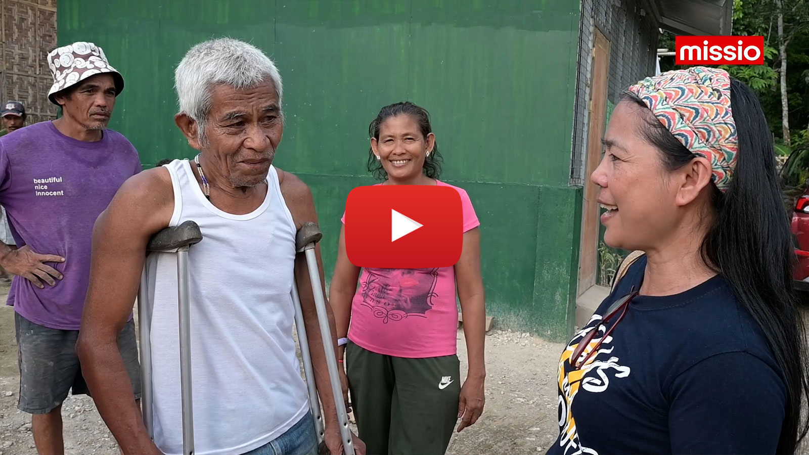 missio-Projektpartnerin Jocelyn Aquiatan (rechts im Bild) unterhält sich auf der Insel Mindanao vor einem grünen Wellblechgebäude mit einem Mann mit Krücken. Hinter ihm stehen eine lächelnde Frau und ein Mann.
