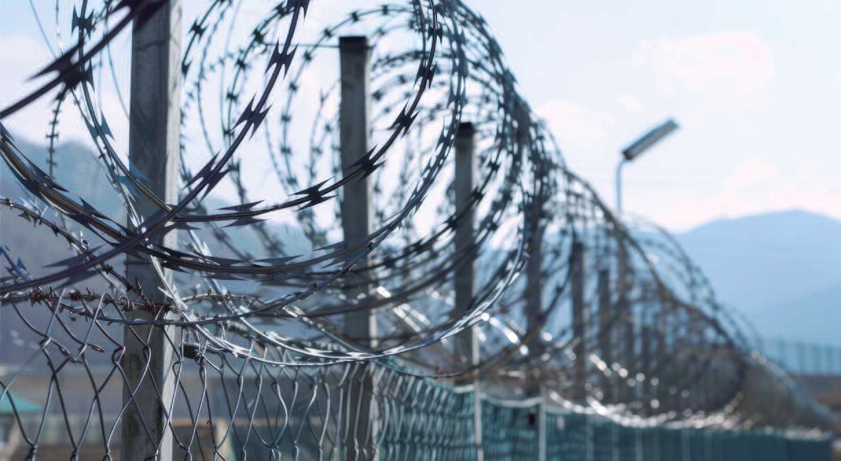 Maschendrahtzaun mit Stacheldraht, dahinter die Silhouette der Berge.