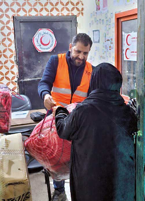 Ein Mann mit orangener Warnweste gibt einer Frau mit Kopftuch eine Plastiktüte mit Decken darin.