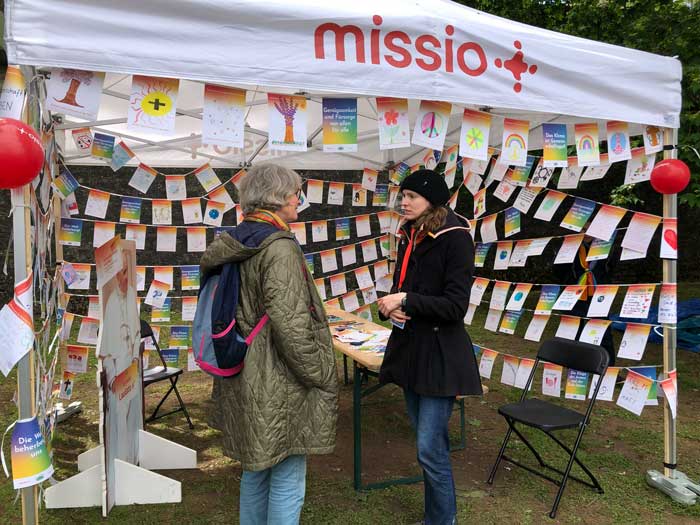 missio-Stand am Katholikentag 2026 in Würzburg mit vielen beschrifteten und bemalten Klimawimpeln. Davor eine missio-Mitarbeiterin im Gespräch mit einer Katholikentagsbesucherin.