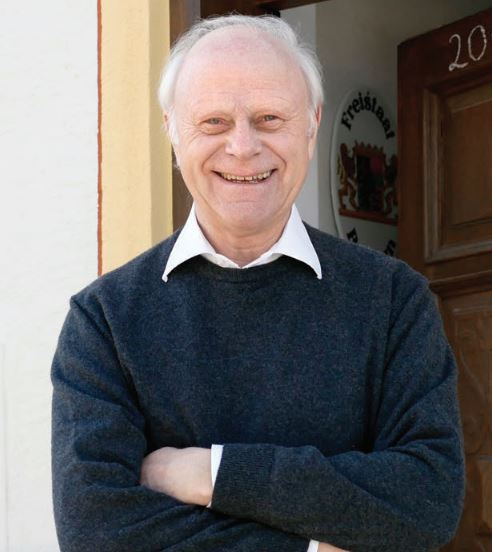 Porträt des Testamentgebers Bernhard Mooser. Erhält die Arme vor der Brust verschränkt und lacht entspannt und freudig in die Kamera.