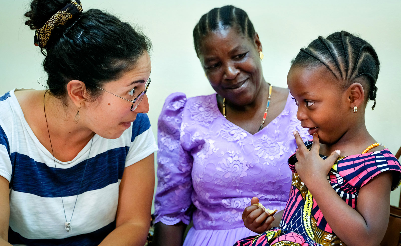 Eine Mitarbeiterin von Simama und ein Mädchen mit Behinderung schauen sich in die Augen. Die Mutter beobachtet im Hintergrund ihre Tochter.
