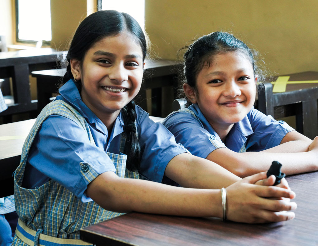 Zwei Schülerinnen in hellblauen Schuluniformen sitzen auf ihrem Platz im Klassenzimmer und lächeln in die Kamera.