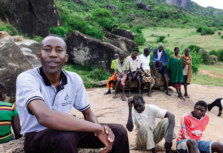 Ein junger Priesterseminarist sitzt im ländlichen Gebiet in Uganda auf einem Felsen. Im Hintergrund sind Gemeindemitglieder zu sehen.