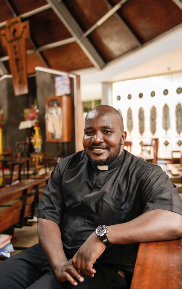 Der Priester Steven Kulana aus Uganda sitzt in einer Kirchenbank und lächelt in die Kamera.
