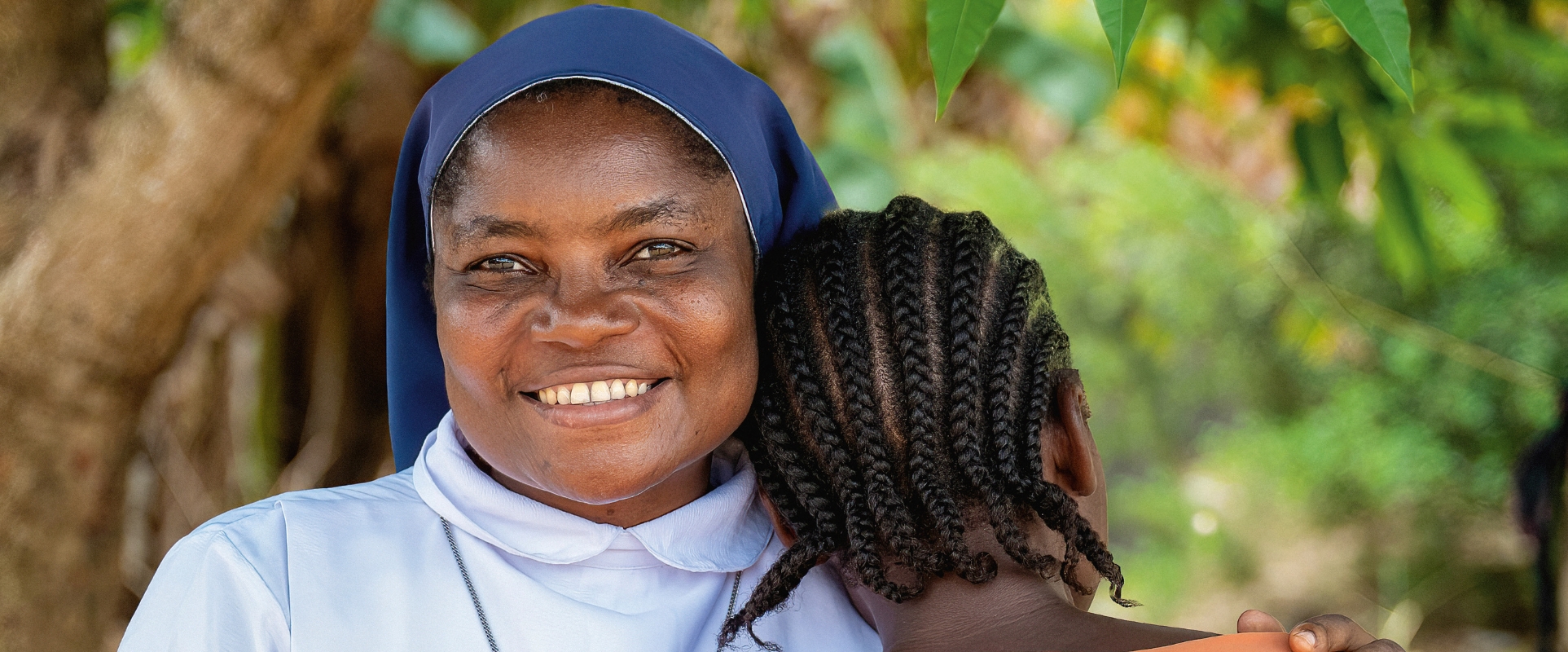 Afrikatag 2027: Eine Ordensschwester mit blauem Schleier und weißem Gewand aus Sierra Leone legt schützend ihre Hände auf die Schultern einer Frau in orangefarbener Kleidung, die dem Betrachter den Rücken zuwendet und ihre Wange an die linke Schulter der Ordensfrau lehnt.