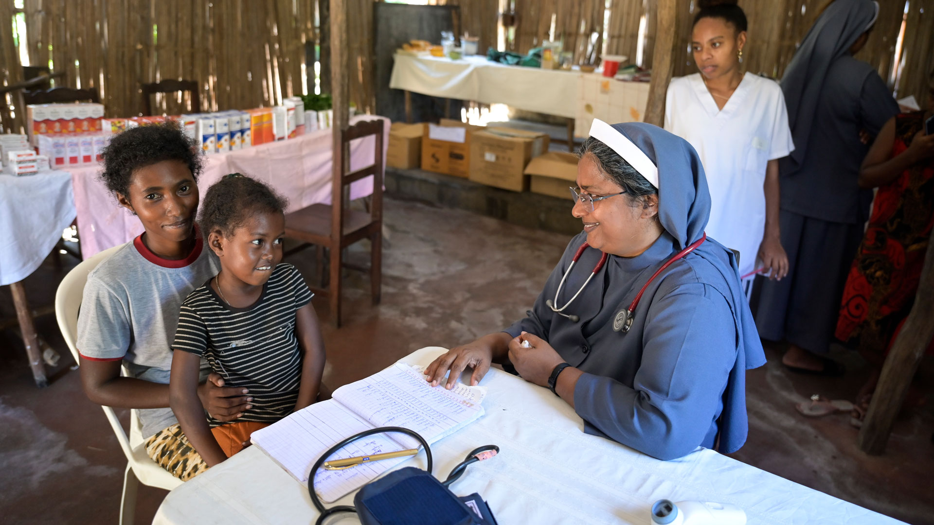 Blick in eine mobile Klinik auf Madagaskar: Sr. Jain Mary am Tisch mit ihrer jungen Patientin Mira, die auf dem Schoß ihrer Mutter sitzt