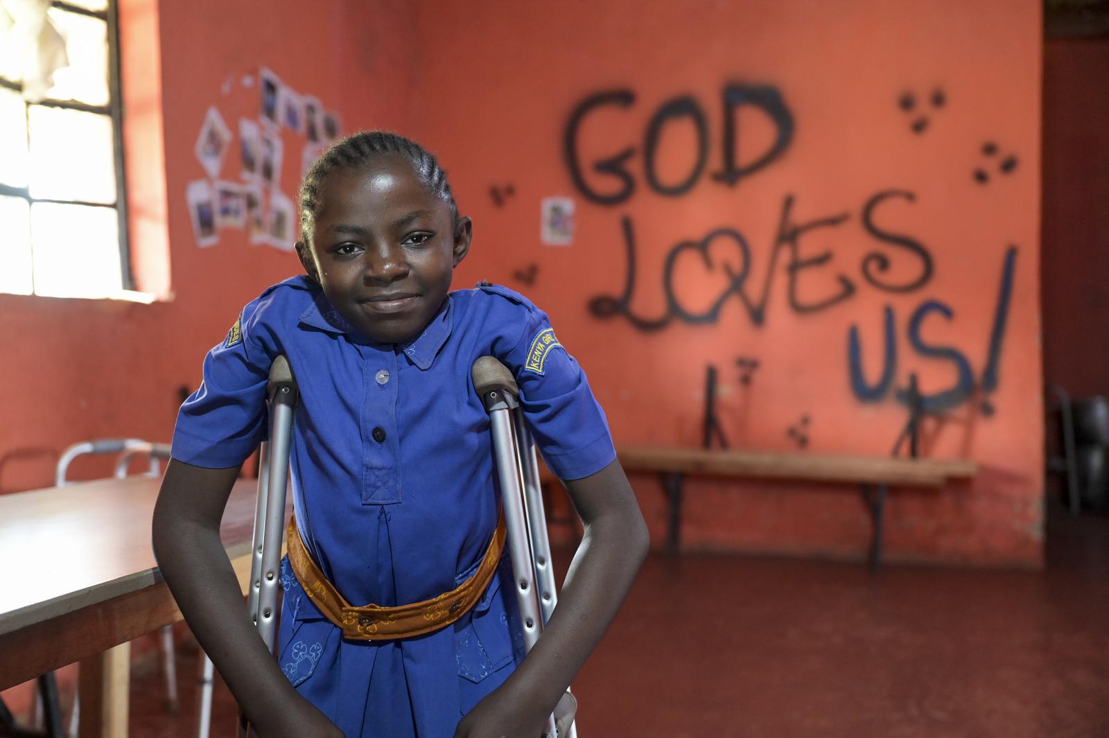 Esther, eine junge Kenianerin,  steht auf ihre Krücken gelehnt in einem Klassenzimmer. Im Hintergrund steht an der Wand: "God loves us".
