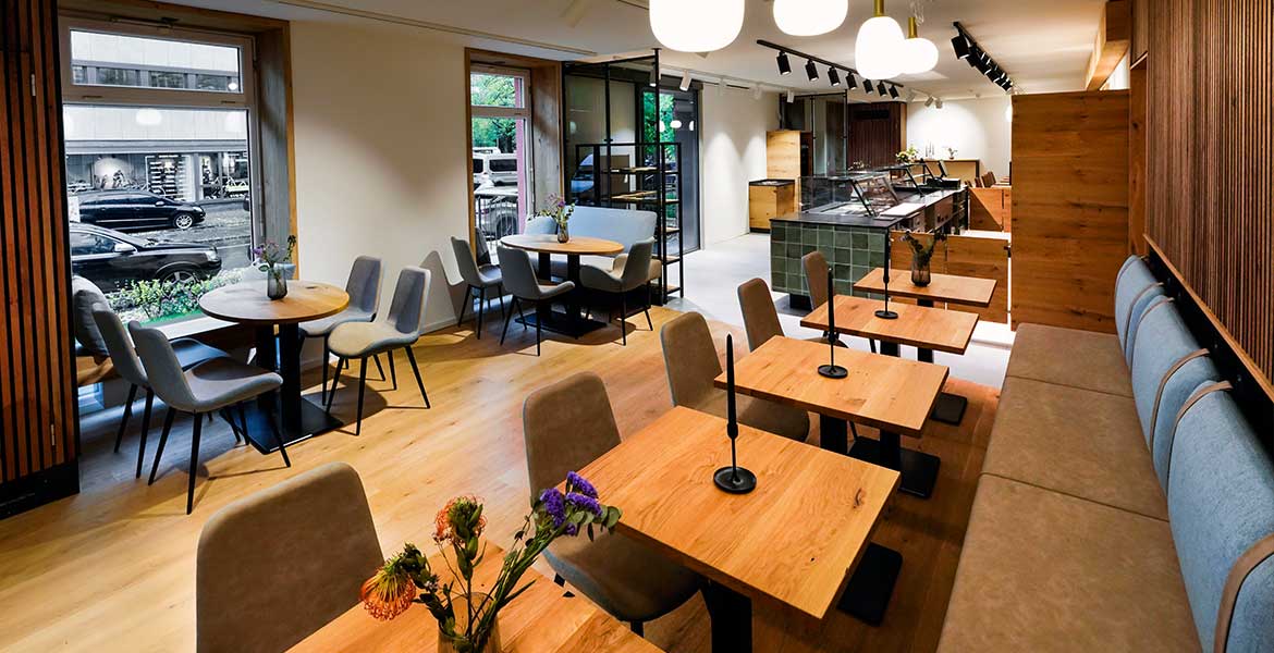 Innenansicht der Cafébar Conviva im Erdgeschoss bei missio München. Blick durch den langegezogenen Raum über die Sitzgruppen in Richtung der Fenster zur Straße.