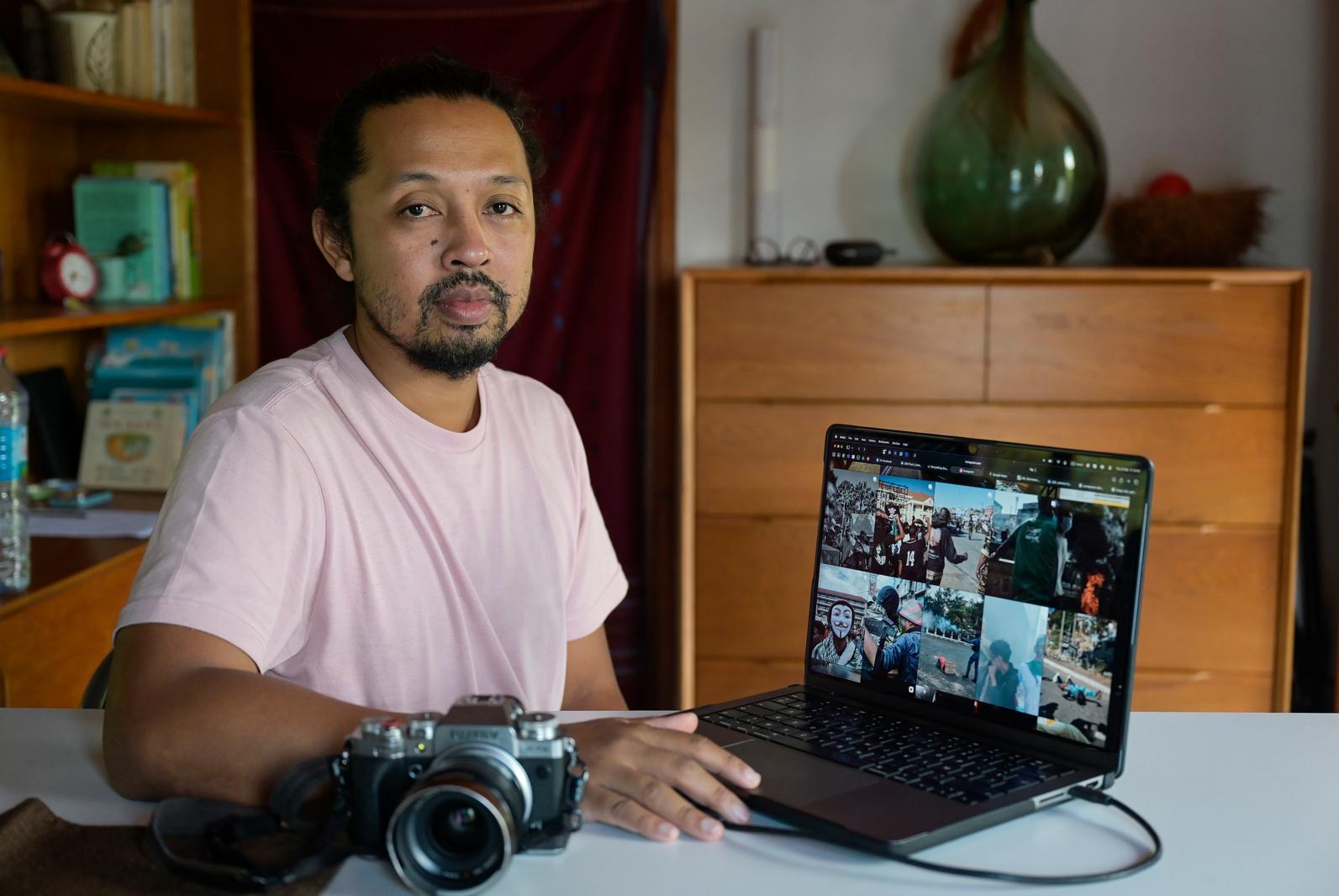 Ein Mann mit zurückgebundenen Haaren und Bart sitzt in einem Raum, in dem ein Regal und eine Kommode aus Holz zu sehen sind. Er trägt ein rosa T-Shirt und hat vor sich eine Kamera liegen. Auf dem Tisch steht sein geöffneter Laptop, leicht in Richtung des Betrachters gedreht, auf dem eine Vorschau verschiedener Bilder von den Protesten zu sehen sind.