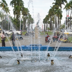 Im Vordergrund ein Springbrunnen, hinter der Fontäne ist ein vielbefahrener Platz zu erkennen. In der MItte eine Statue, rechts und links eine lange Reihe Palmen und dazwischen Fahnenmasten mit der madagasischen Flagge.