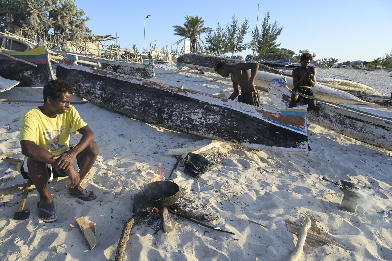 Drei Fischer neben ihren Booten am Strand. Einer kocht etwas auf einer Feuerstelle.