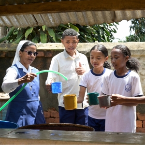 Schwester Rolandine steht mit vier Schülerinnen hinter einem Brunnen in einer Schule in Analavory in Madagaskar und gießt mit einem Schlauch frisches Wasser in die Becher der Kinder.
