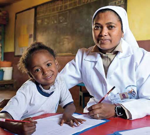 Ein Mädchen sitzt in einem Klassenzimmer in Madagaskar und schreibt in ein Heft. Eine Ordensschwester hilft ihr. Beide lächeln in die Kamera.