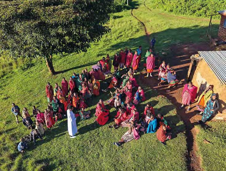 Von oben ist der Vorplatz einer Boma im Ngorongoro-Naturschutzgebiet in Tansania zu sehen. Ein weiß gewandeter Katechist spricht zu einer Gruppe von buntgekleideten Frauen und Kindern.