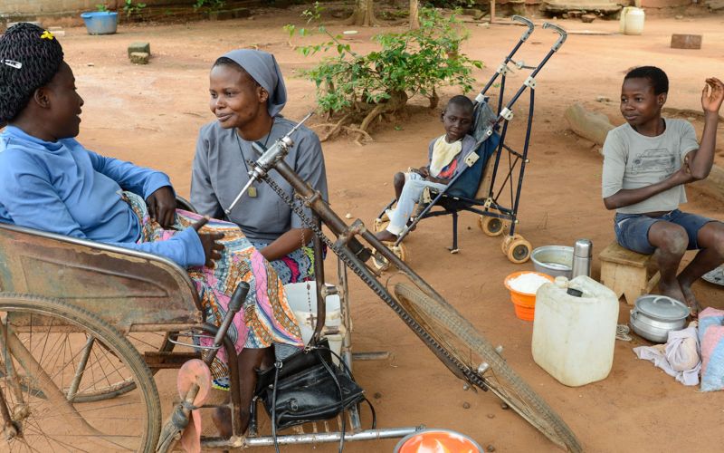 Eine Ordensfrau sitzt in einem Hof bei einer Frau im Rollstuhl und hört ihr zu. Im Hintergrund ist ein Kind in einem Buggy zu sehen und ein größeres Kind sitzt an einer Kochstelle.