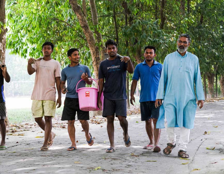 Der Priester Anand (ganz rechts im Bild) läuft mit vier jungen Männern, die Spaten, Schaufeln und zwei Eimer tragen, einen Weg entlang und auf auf die Kamera zu.
