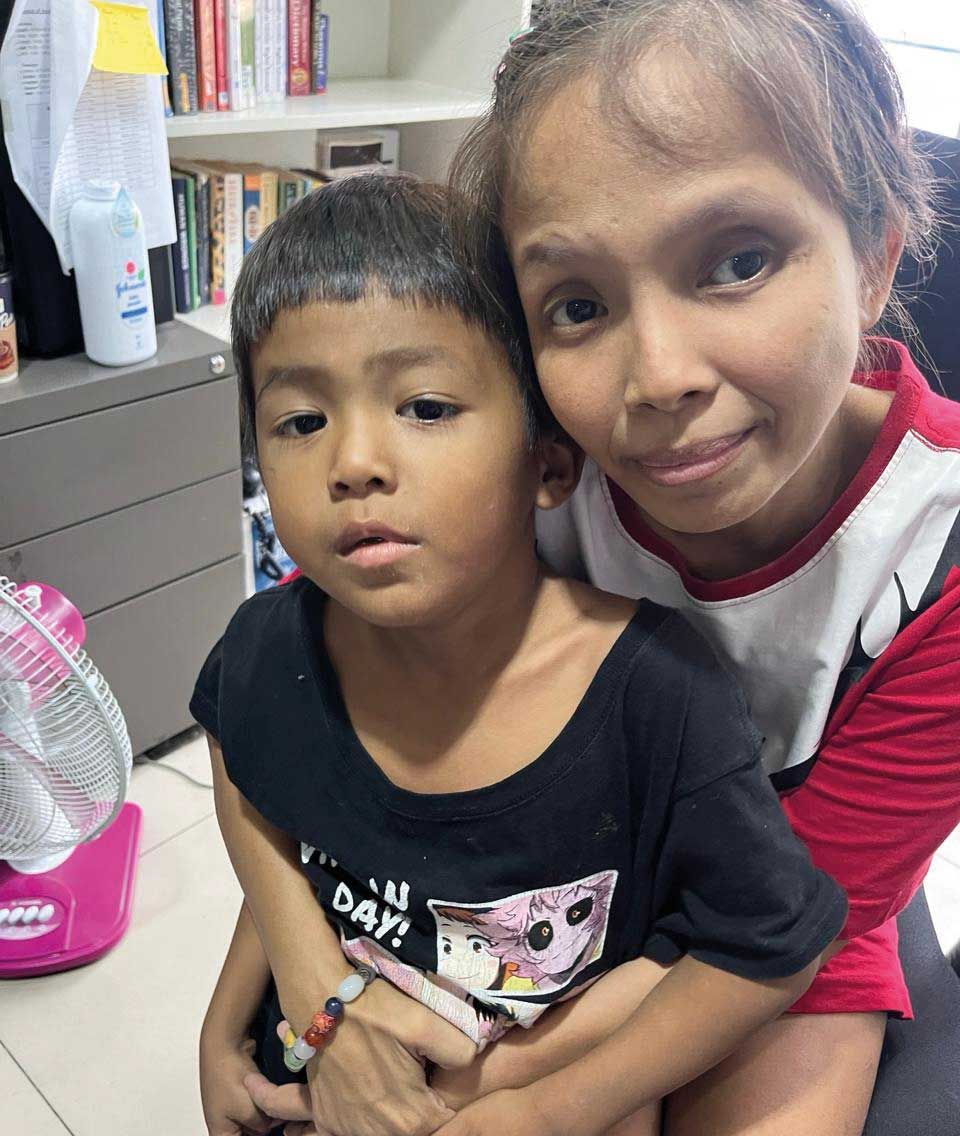 Drei Frauen und ein Mann aus der indigenen Bevölkerungsgruppe in Mindanau auf den Pilippinen sitzen in bunten, folkloristischen Kleidern auf einer Holzbank vor einem Gebäude aus Bambus. Ein kleines Kind steht neben einer der sitzenden Frauen und sieht ebenfalls in die Kamera.