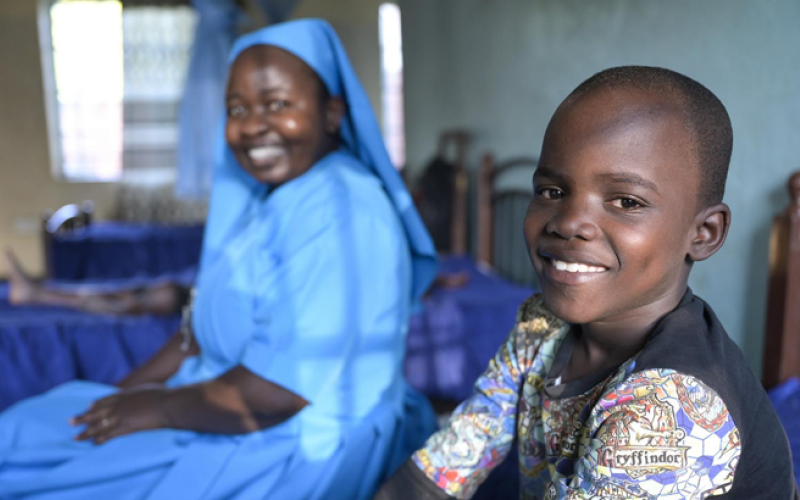 Ein Junge sitzt auf einem Bett im Kinderschutzzentrum Lusubilo in Karonga/Malawi und lacht in die Kamera. Im Hintergrund sitzt eine Ordensschwester und blickt ebenfalls lächelnd in die Kamera.