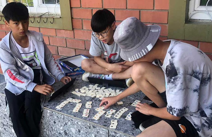 Drei Jungen sitzen auf einer Steinbank für einem Backsteingebäude in der Mongolei und spielen mit einer Art Kartenspiel.