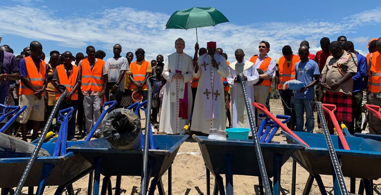 Alles ist vorbereitet für den Spatenstich zum Kirchenbau für den Bildungscampus am Turkana-See in Kenia. Im Vordergrund des Bildes sind Schubkarren und Spaten zu sehen. Dahinter eine ganze Reihe von Männern mit orangener Warnweste. In der Mitte stehen von links nach rechts missio-Präsident Monsignore Wolfgang Huber, der örtliche Bischof, ein weiterer Geistlicher sowie Ludwig Prinz von Bayern.