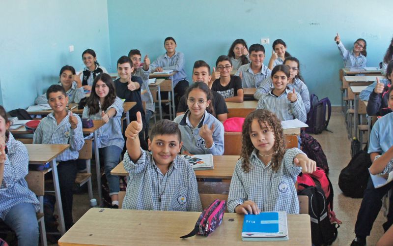 Schulkinder aus dem Libanon sitzen in hellblauer Schuluniform in einem Klassenzimmer an Schultischen. Sie lachen in die Kamera, einige von ihnen strecken ihren Daumen nach oben.