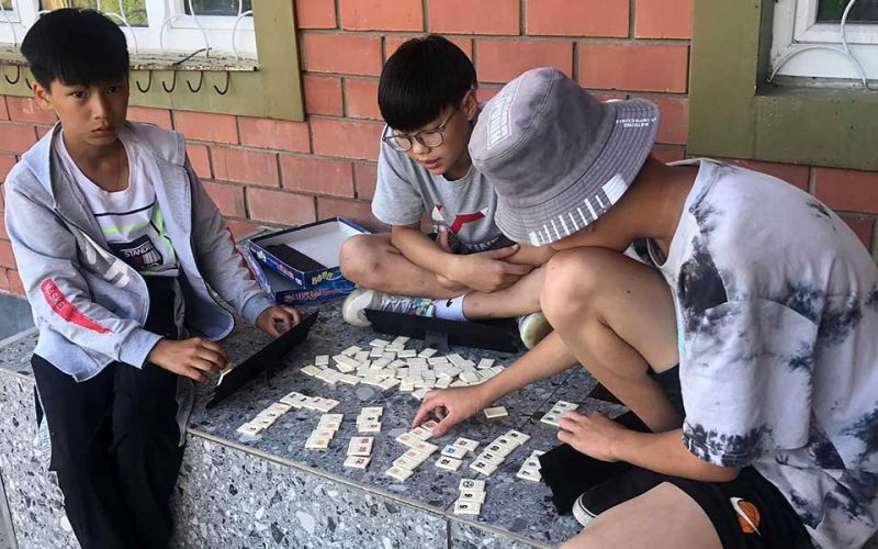 Drei Jungen sitzen auf einer Steinbank für einem Backsteingebäude in der Mongolei und spielen mit einer Art Kartenspiel.