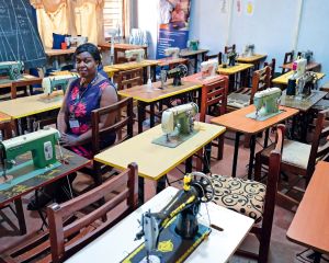 Eine Frau sitzt allein im Schulungsraum, vorne eine Tafel mit Schnittmustern, auf den Tischen stehen Nähmaschinen. Davor je ein leerer Stuhl.