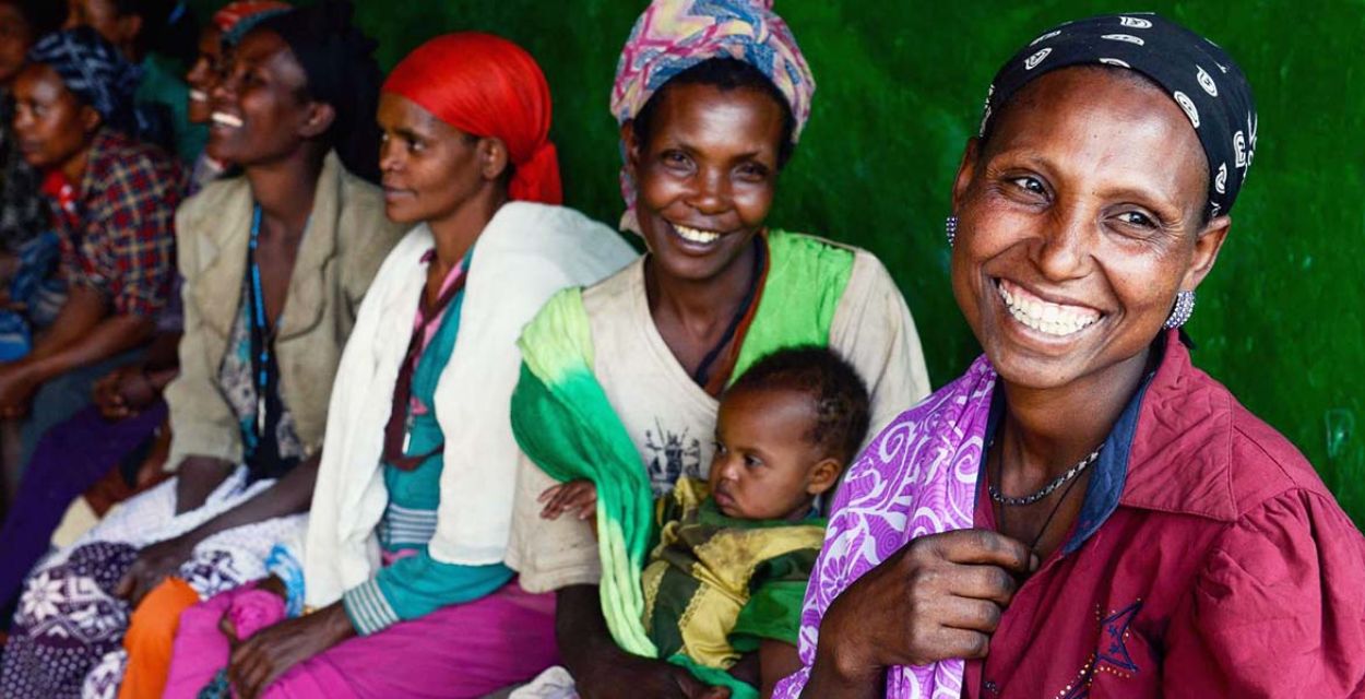 Frauen in Äthiopien in bunten Kleidern sitzen auf einer Bank vor einer grünen Wand. Eine von ihnen hält ein Baby auf dem Schoß. Foto: Jörg Böthling/missio München