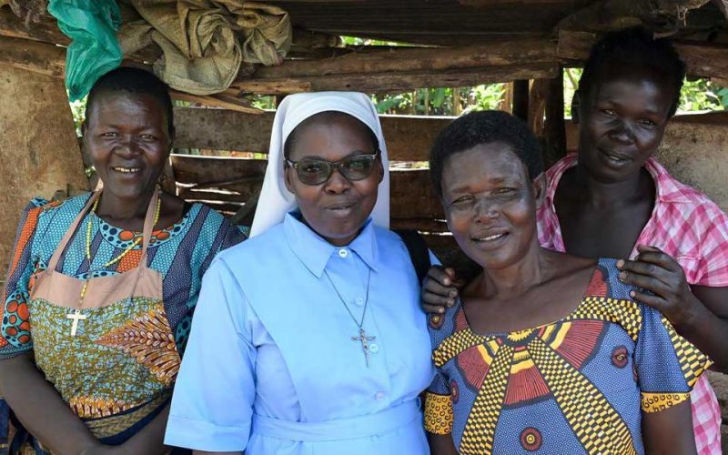 Vier Frauen aus Uganda stehen unter vor einer Bretterwand mit Dach und lächeln in die Kamera. Die zweite von links ist eine Ordensschwester.