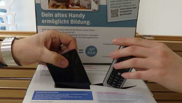Zwei Hände werfen von links und rechts ausrangierte Handys in eine Spendenbox.