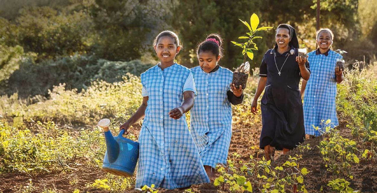 Eine Ordensfrau und drei Mädchen laufen in Madagaskar über ein Feld auf die Kamera zu. Das erste Mädchen trägt in der rechten Hand eine Gießkanne, das hinter ihr laufende Mädchen, die Ordensschwester dahinter und das letzte Mädchen tragen jeweils einen Pflanzensetzling in ihren linken Händen. Alle vier lächeln.