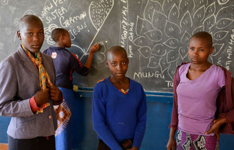 Vier junge Frauen stehen vor einer Schultafel. Drei von ihnen schauen in die Kamera, eine steht hinten an der Tafel und schreibt etwas mit Kreide darauf.