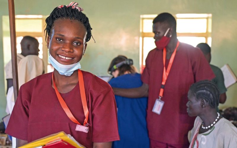 Links im Bild: die angehende Hebamme Edia in ihrer bordeauxroten Arbeitskleidung. Sie hat die OP-Maske heruntergezogen und lächelt. Im Arm hält sie Patientenakten. Im Hintergrund rechts von Edia: ein anderer Angestellter des Krankenhauses sowie weitere Personen, vielleicht Patienten.