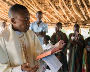 Ein Priester steht in Uganda unter einer Überdachung aus Holz und Lehm und liest aus einer aufgeschlagenen Bibel. Im Hinjtergrund sieht man betende Gläubige.
