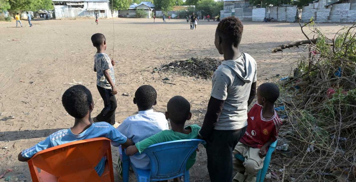 Vier Kinder sitzen im Halbschatten auf Plastikstühlen. Zwei weitere stehen. Sie wenden der Kamera den Rücken zu und blicken auf einen weitläufigen, sandigen Platz.