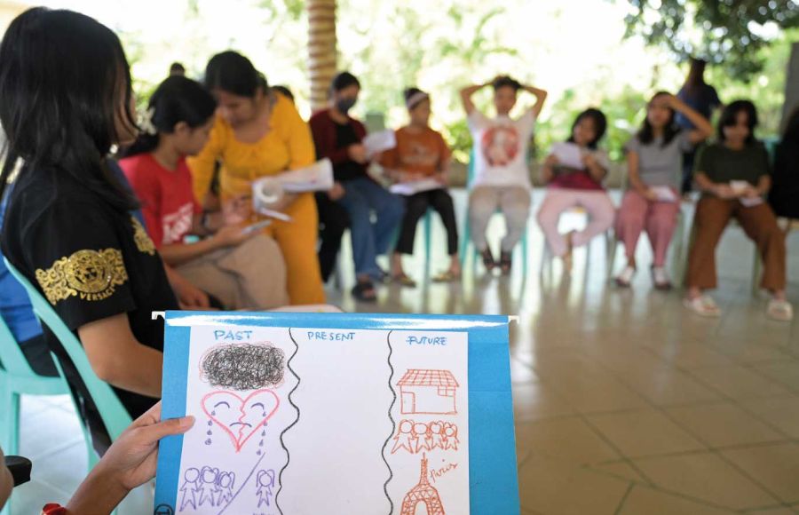 Mädchen sitzen im Hintergrund im Schutzzentrum Hablondawani in einem Kreis. Im Vordergrund ist eine Hand zu sehen, die eine Zeichnung in die Höhe hält.