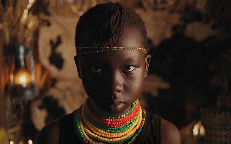 Zu sehen ist die Hauptdarstellerin des Kinofilms "Nawi". Das junge Mädchen trägt viele bunte Ketten um den Hals und zwei Ketten um den Kopf. Sie blickt den Betrachter unverwandt und leicht von unten an. Hinter ihr hängt eine Gasterne die das Fell im Inneren der Hütte beleuchtet.