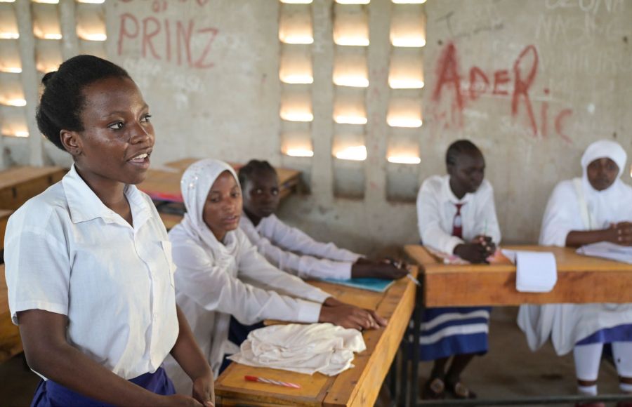 Schülerinnen an der Blue State Secondary des JRS in Kenia sitzen in einem Klassenzimmer. Ein Mädchen im Vordergrund steht und spricht in Richtung Tafel nach vorne.
