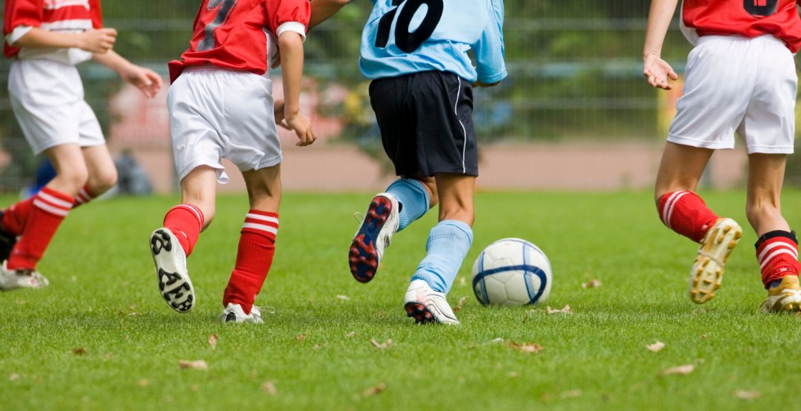 Auf einem Rasenplatz liegt ein Fußball. Vier Jungen bewegen sich auf den Ball zu. Zu sehen sind nur Beine und Rumpf. Drei Jungs tragen ein rotes Trikot mit weißer Hose, ein weiterer Junge ein blaues Trikot mit schwarzer Hose.