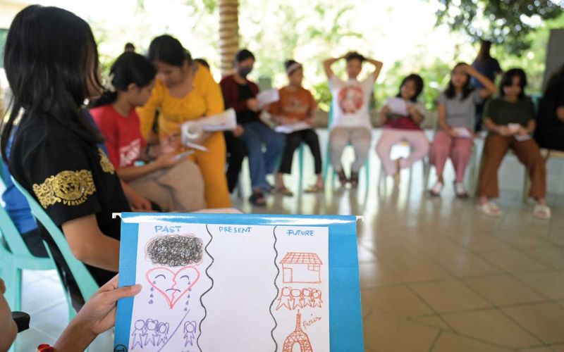 Mädchen sitzen im Hintergrund im Schutzzentrum Hablondawani in einem Kreis. Im Vordergrund ist eine Hand zu sehen, die eine Zeichnung in die Höhe hält.