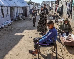 Geflüchtete Palästinenser aus dem Gazastreifen sitzen und stehen auf einer Lehmstraße. Links sind Zelte zu sehen, rechts aufgehängte Wäsche an einer Mauer.